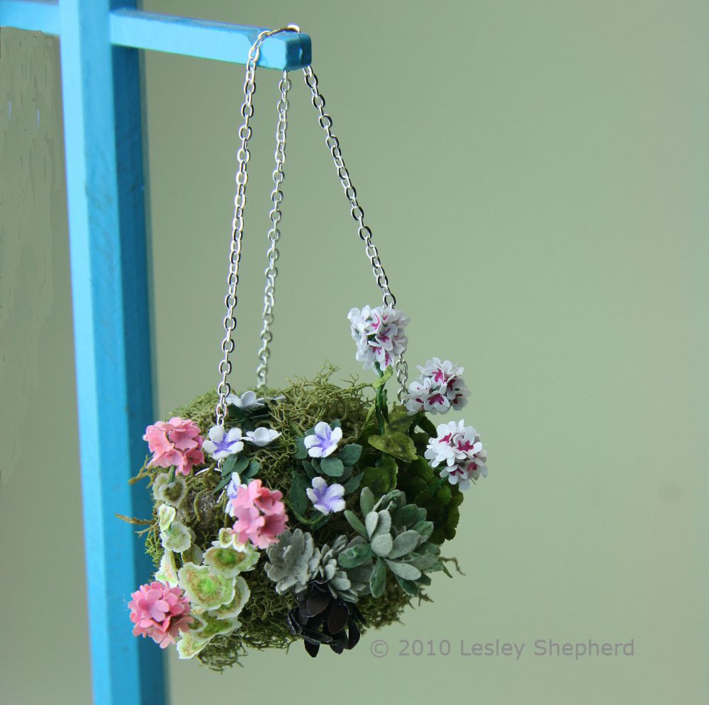 A dolls house scale planted moss hanging basket hung from a chain on a lampstand.