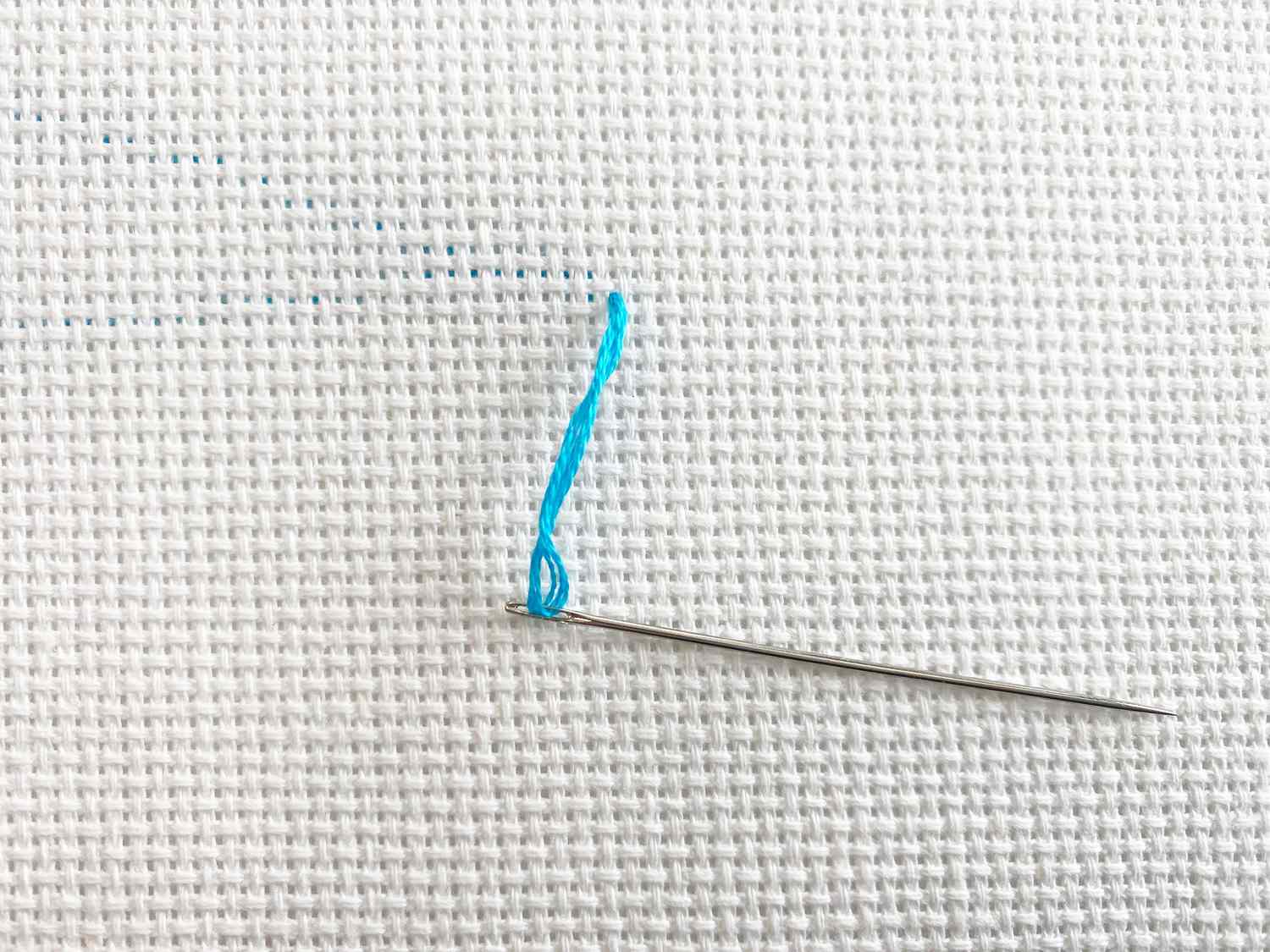 A close-up of blue thread in a needle with Aida cloth in the background