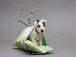 A dolls house scale Jack Russell Terrier tucked into the cushion on his dog bed.