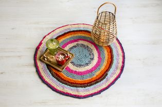 Colorful crochet rug from cut repurposed T-shirts on white wooden home floor with wood lantern, candle burning. Cup on hot chocolate wit marshmallows on tray. Flat lay view.