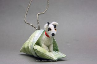 A dolls house scale Jack Russell Terrier tucked into the cushion on his dog bed.