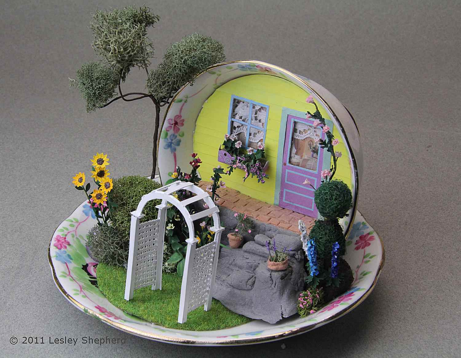 Quarter scale garden arbor set in a miniature garden scene in a tea cup.