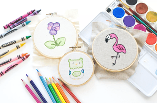 Three embroidery pieces in hoops surrounded by watercolor, crayons, and colored pencils