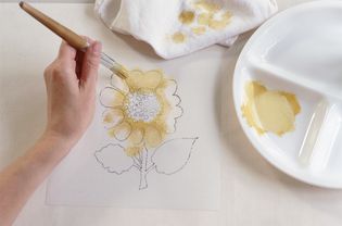 Hand blotting pale yellow paint onto petals of sunflower stencil, painting white fabric below, using wooden-handled paint brush.