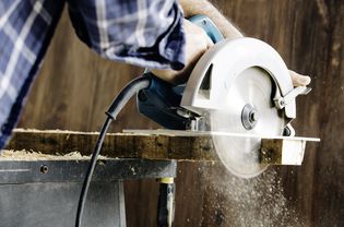 Male carpenter using electric circular saw in home workshop with wood chips flying