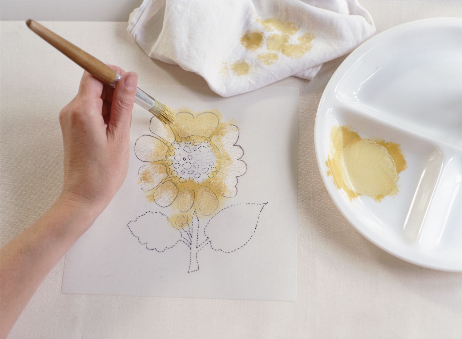 用手在向日葵模板的花瓣上涂上淡黄色的颜料，用木柄画笔在下面涂上白色的织物。