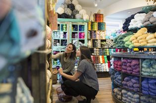 Yarn store owner helping customer look through shelves of colorful yarn.